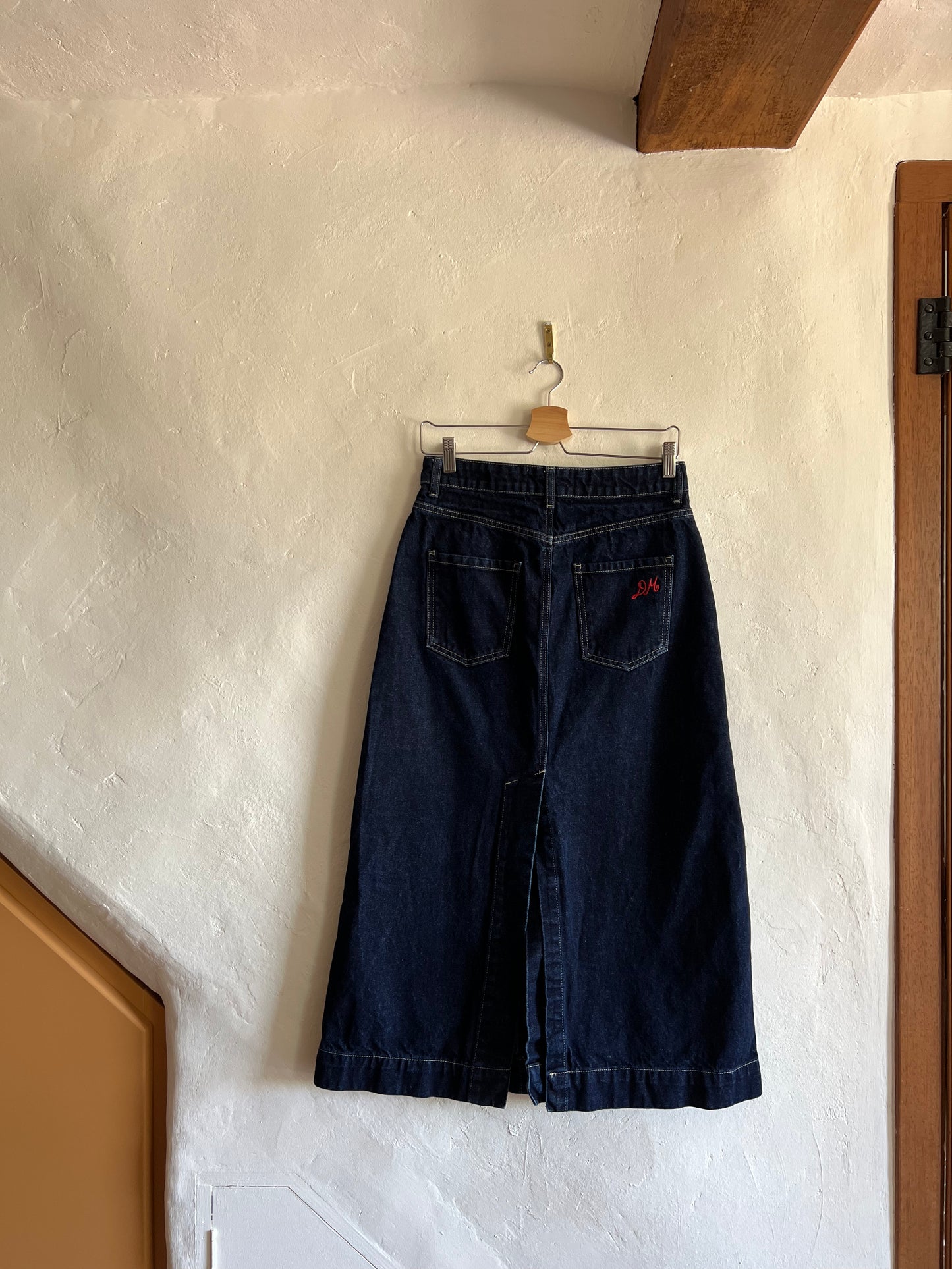 Damson Madder Dark Denim Midi Skirt hanging against a white wall 