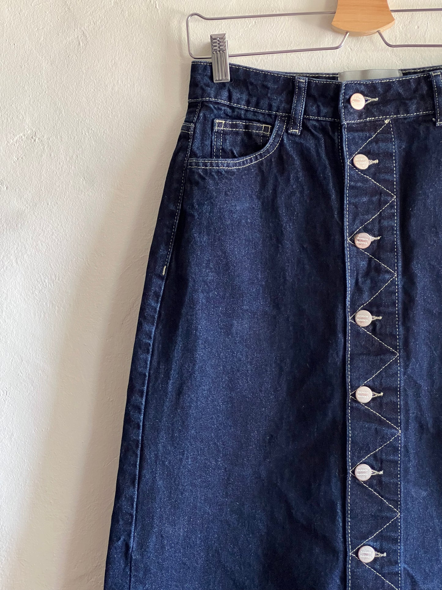 Damson Madder Dark Denim Midi Skirt hanging against a white wall 